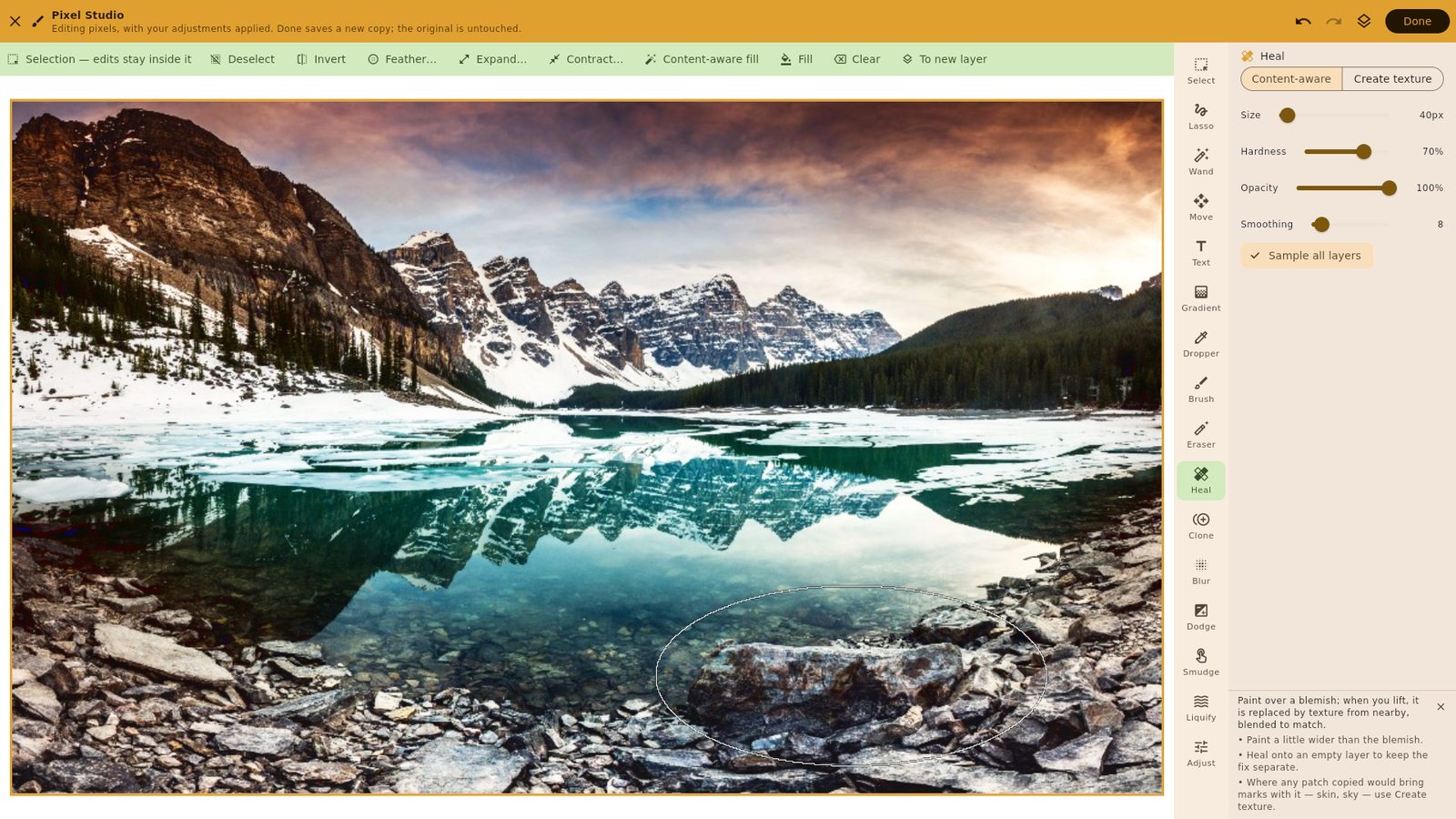 Pixel Studio on a wide window: the amber mode bar, a selection bar with content-aware fill, the photograph framed in amber with an elliptical selection over the foreground rocks, the tool rail and the healing brush's settings
